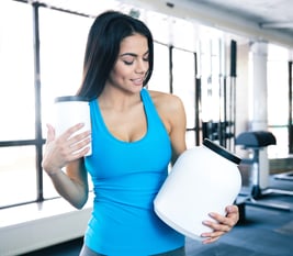 Happy young woman holding plastic container with sports nutrtion at gym and looking on container